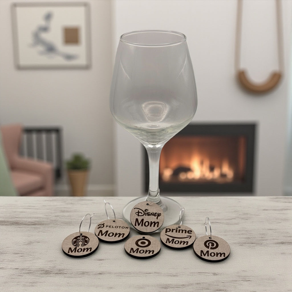 A wine glass by a lit fireplace with a stack of round wooden Mom Life wine charms beside it