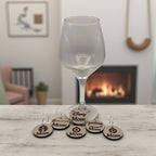 A wine glass by a lit fireplace with a stack of round wooden Mom Life wine charms beside it