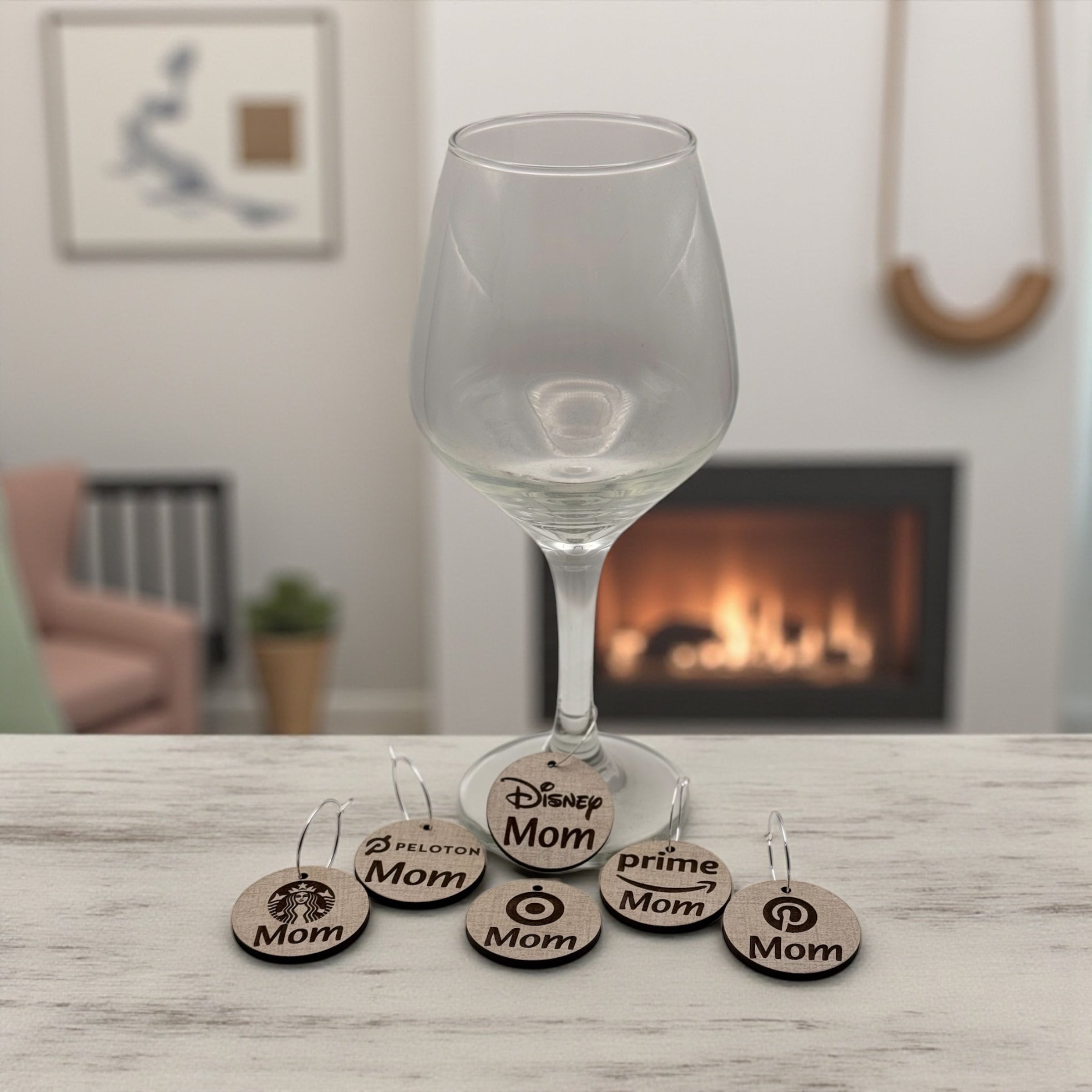 A wine glass by a lit fireplace with a stack of round wooden Mom Life wine charms beside it