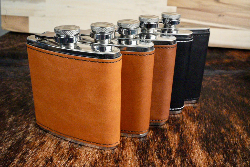 Group of 6 oz. leatherette hip flasks in tan and black, standing on a cowhide surface