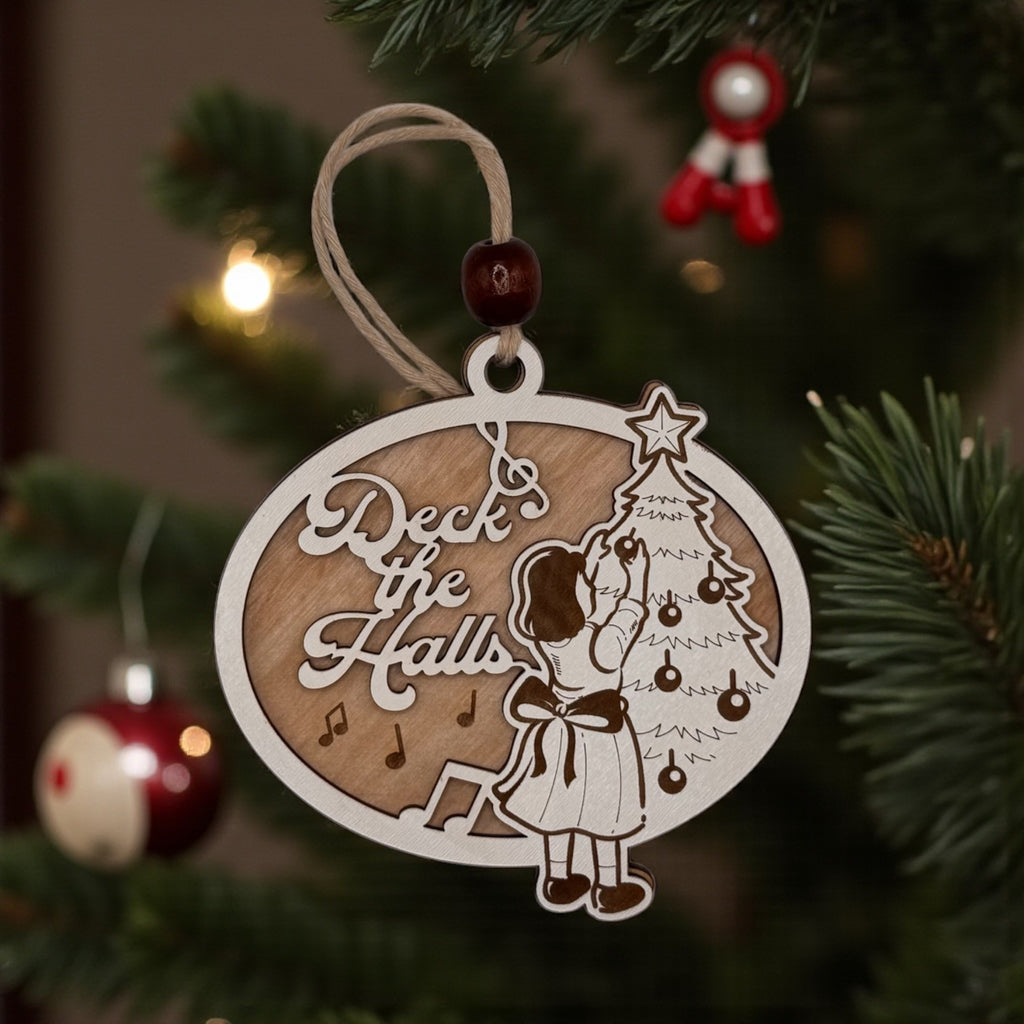 Wooden oval ornament engraved with a Deck the Halls scene of a girl trimming a Christmas tree, hanging on a pine bough with red baubles