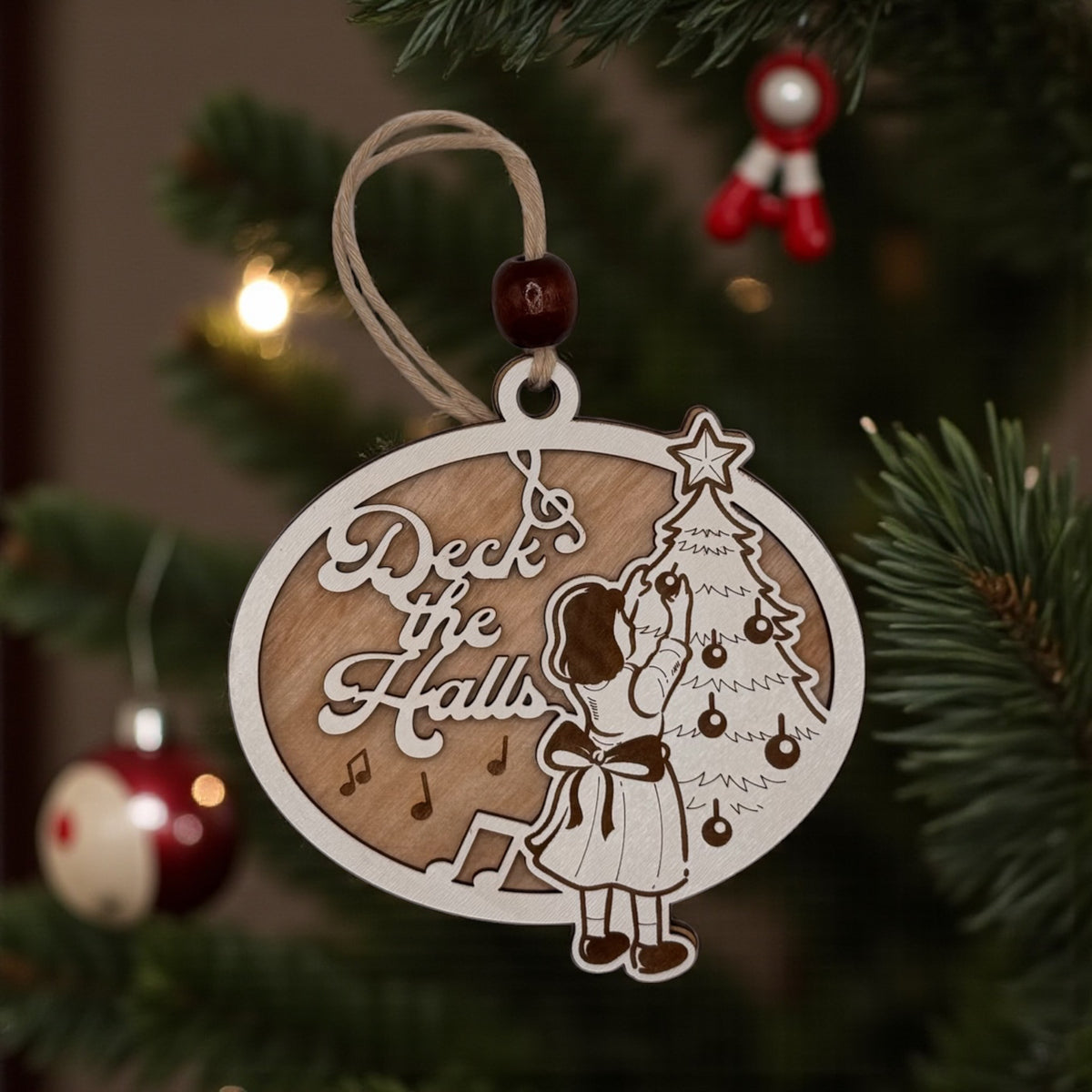Wooden oval ornament engraved with a Deck the Halls scene of a girl trimming a Christmas tree, hanging on a pine bough with red baubles