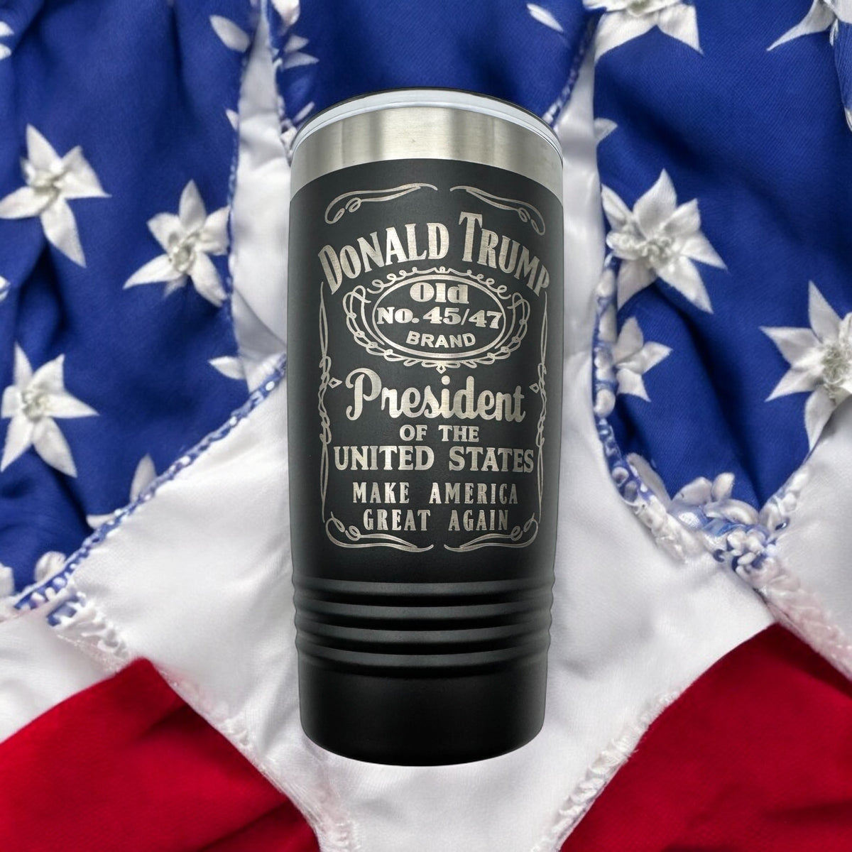 Black tumbler engraved with a whiskey-label-style Donald Trump design, photographed against an American flag