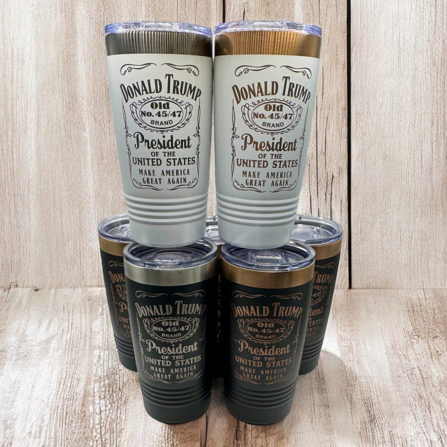 Six engraved tumblers in white, black and graphite standing on a wooden shelf, each carrying the same whiskey-label-style Donald Trump design