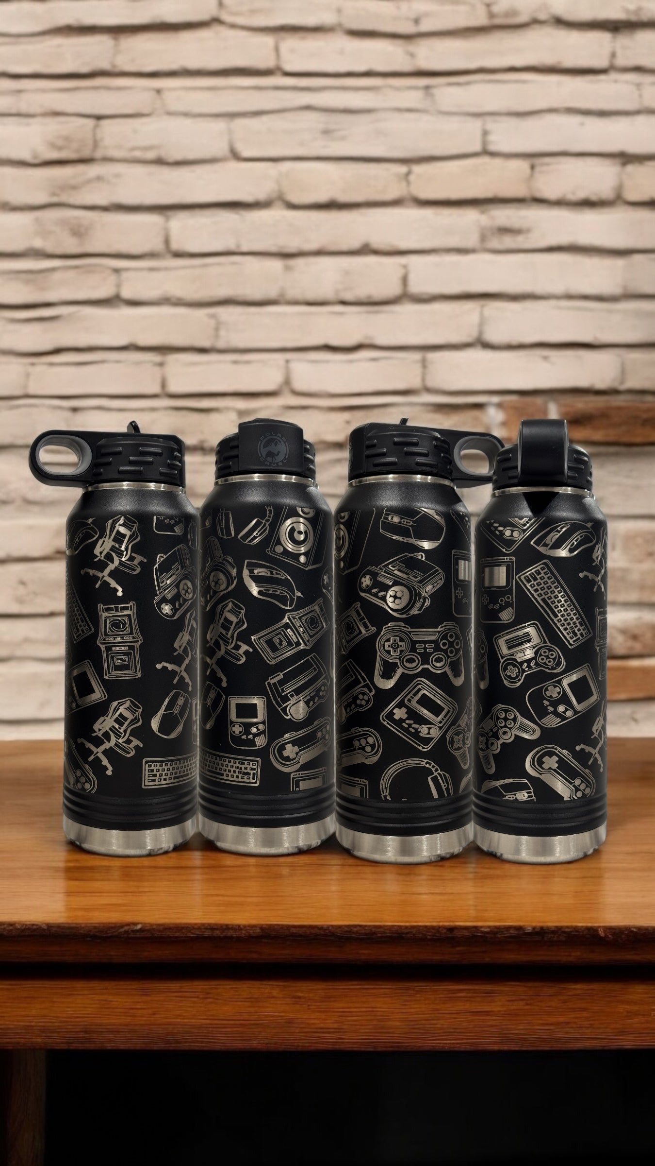 A row of black insulated water bottles engraved with a collage of video game icons, standing on a wooden table against a brick wall
