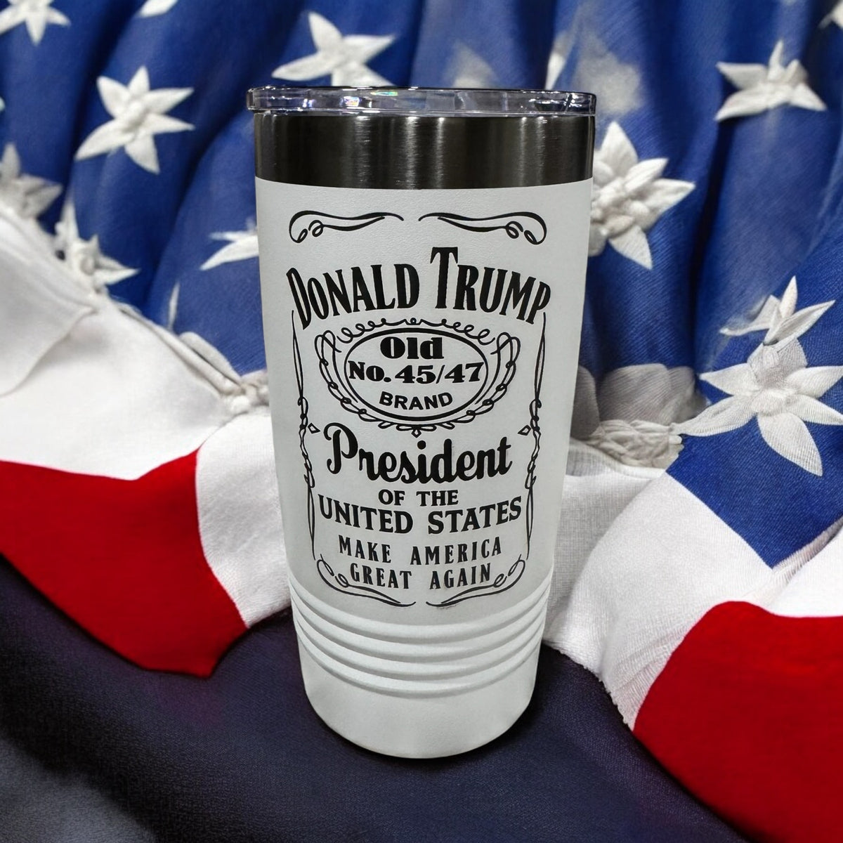White tumbler engraved with a whiskey-label-style Donald Trump design, photographed against an American flag