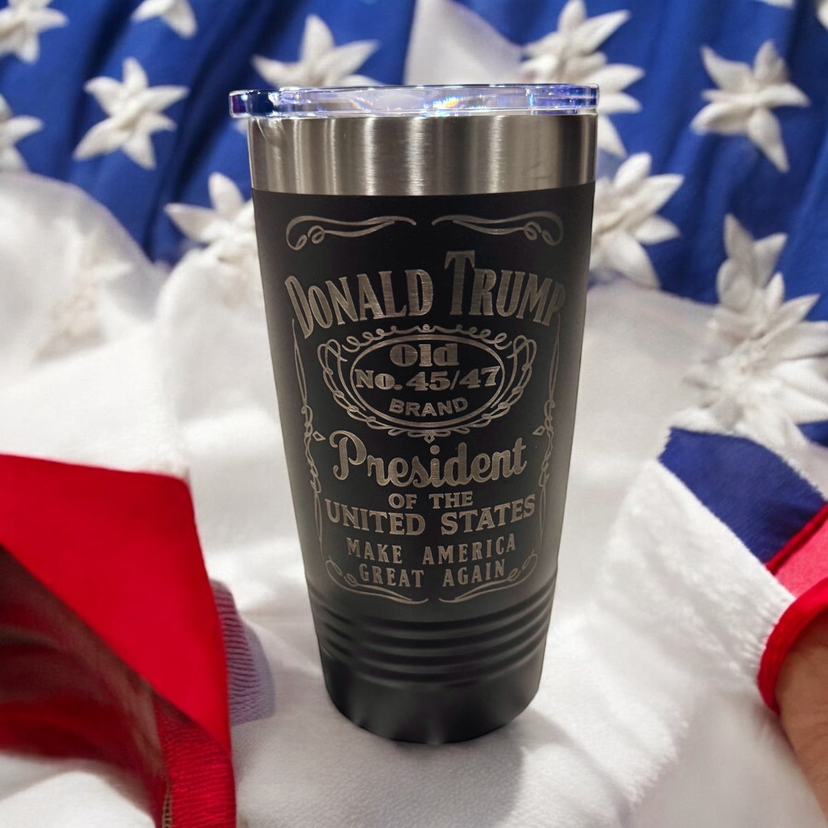 Graphite tumbler engraved with a whiskey-label-style Donald Trump design, photographed against an American flag