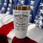 White tumbler with a gold rim, engraved with a whiskey-label-style Donald Trump design, photographed against an American flag