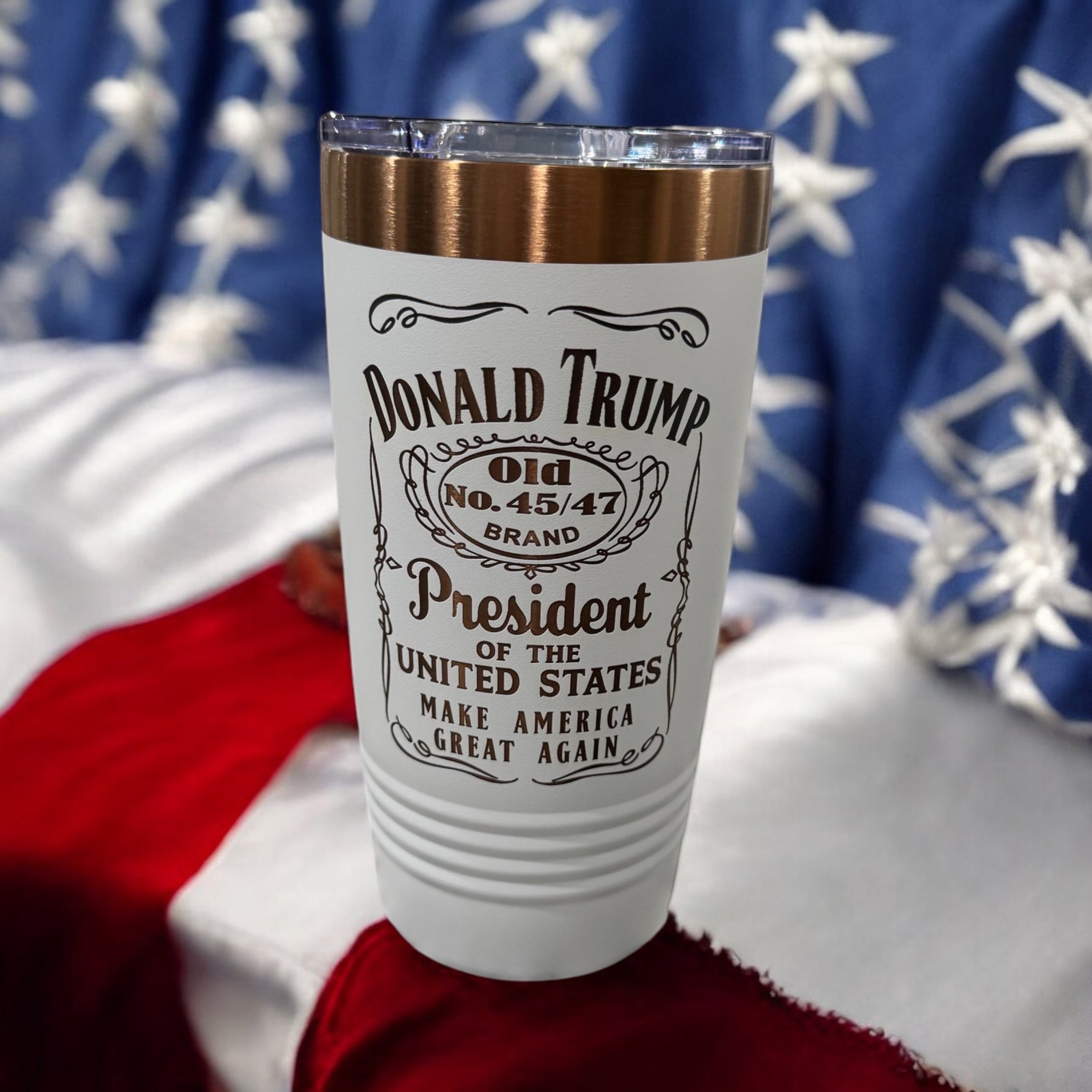 White tumbler with a gold rim, engraved with a whiskey-label-style Donald Trump design, photographed against an American flag