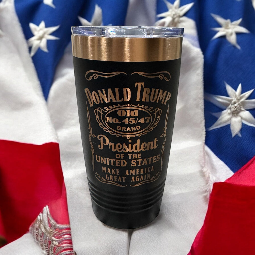 Black tumbler with a gold rim, engraved with a whiskey-label-style Donald Trump design, photographed against an American flag