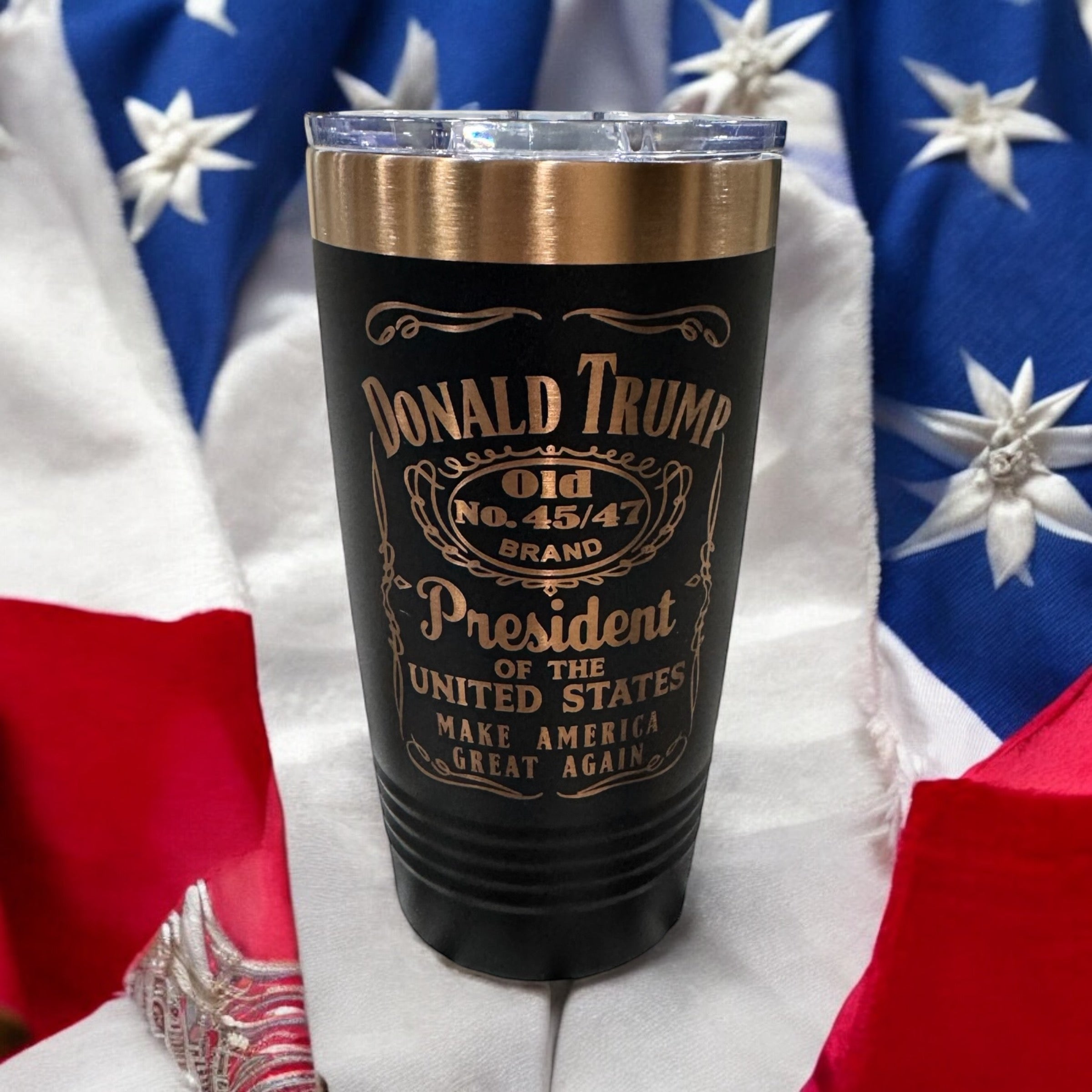 Black tumbler with a gold rim, engraved with a whiskey-label-style Donald Trump design, photographed against an American flag