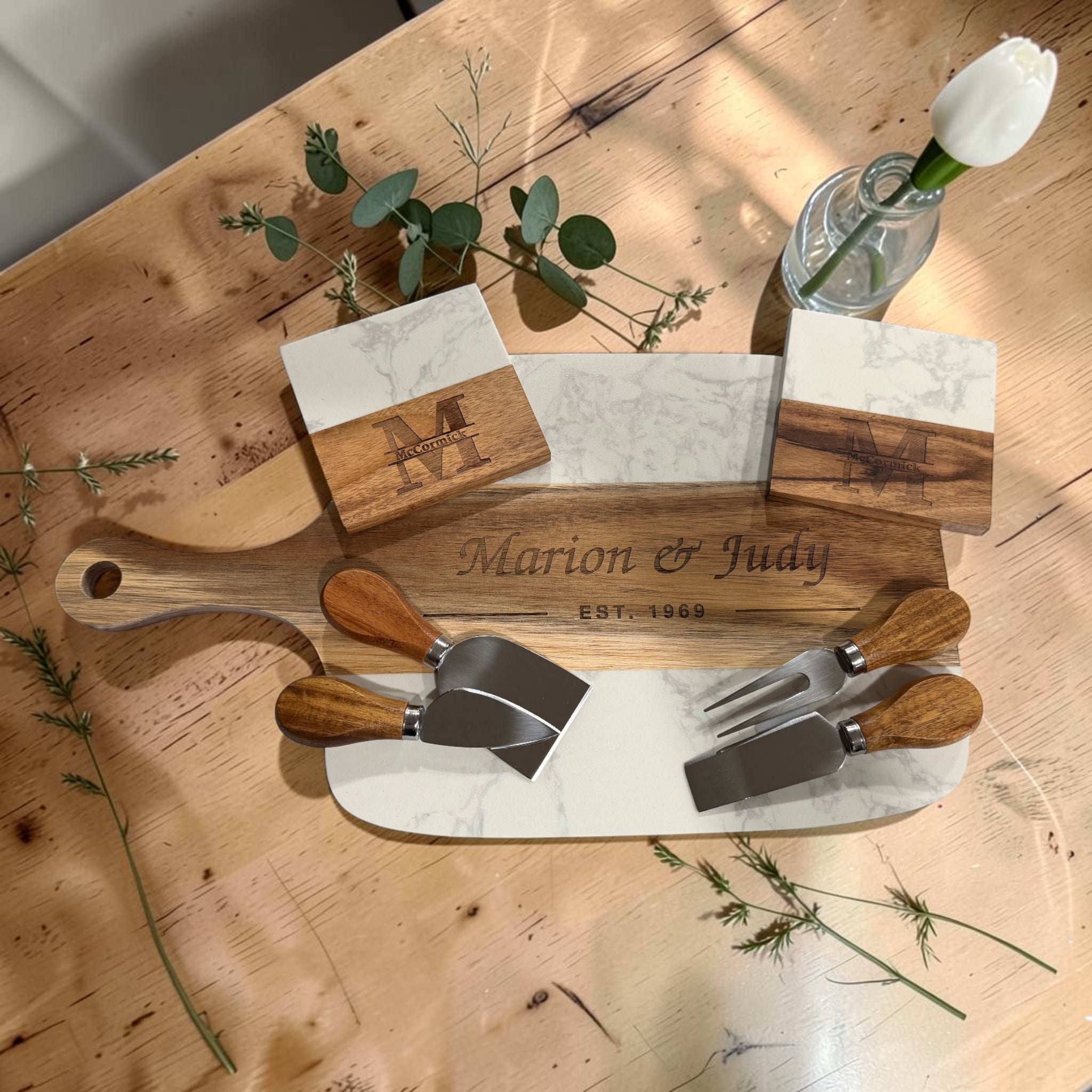 Overhead view of the engraved marble and acacia serving board with three cheese tools and two monogrammed coasters on a rustic wooden table