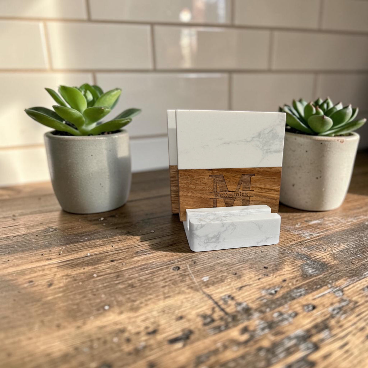 Close-up of marble and acacia coasters engraved with a family name, standing in their matching marble holder between two potted succulents