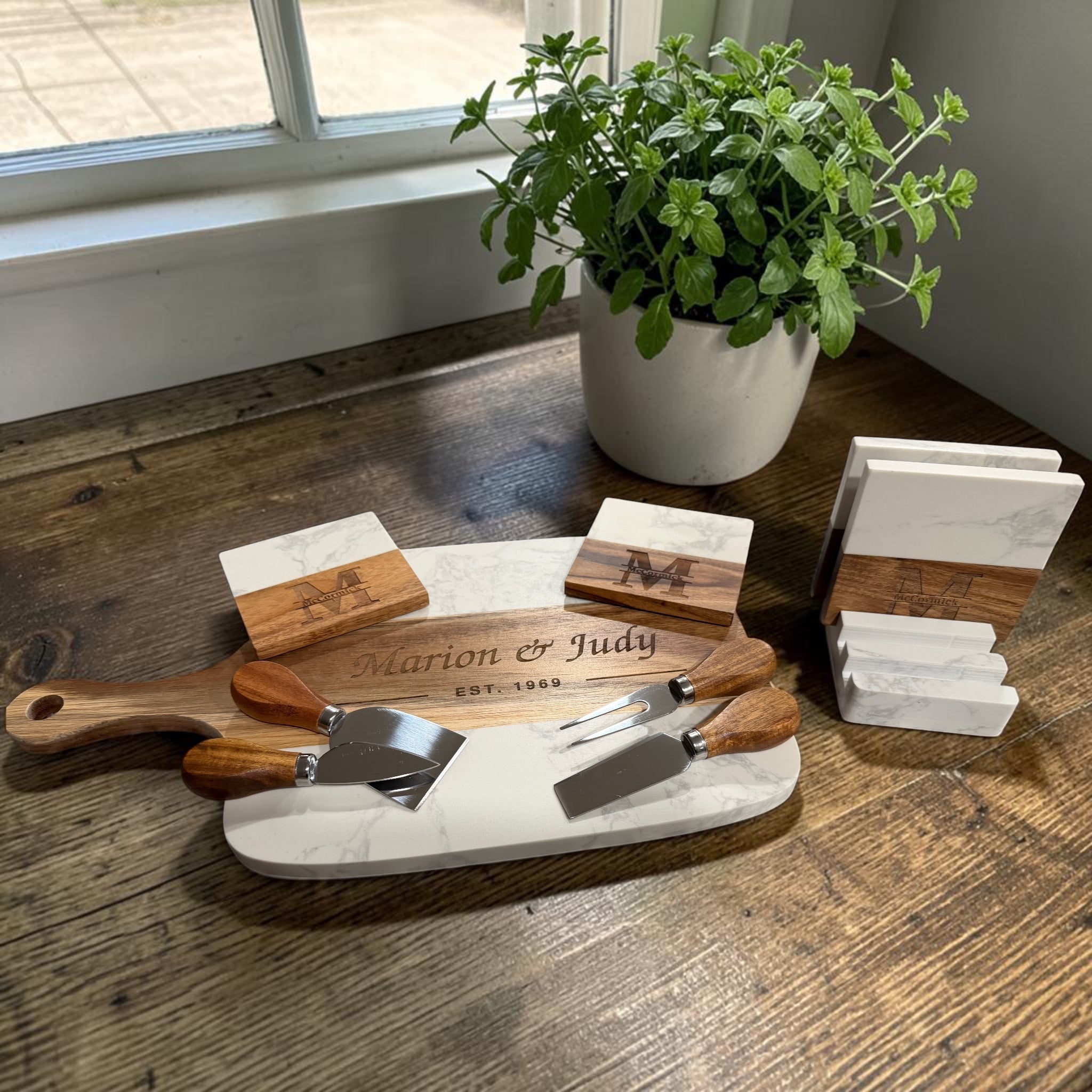 Engraved marble and acacia serving board with three cheese tools, monogrammed coasters and their holder, on a wooden table by a sunlit window