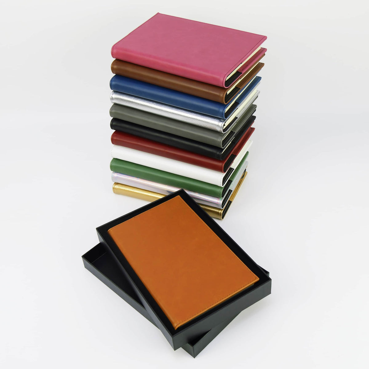 Stack of leatherette journals in every color beside a tan journal resting on its black gift box