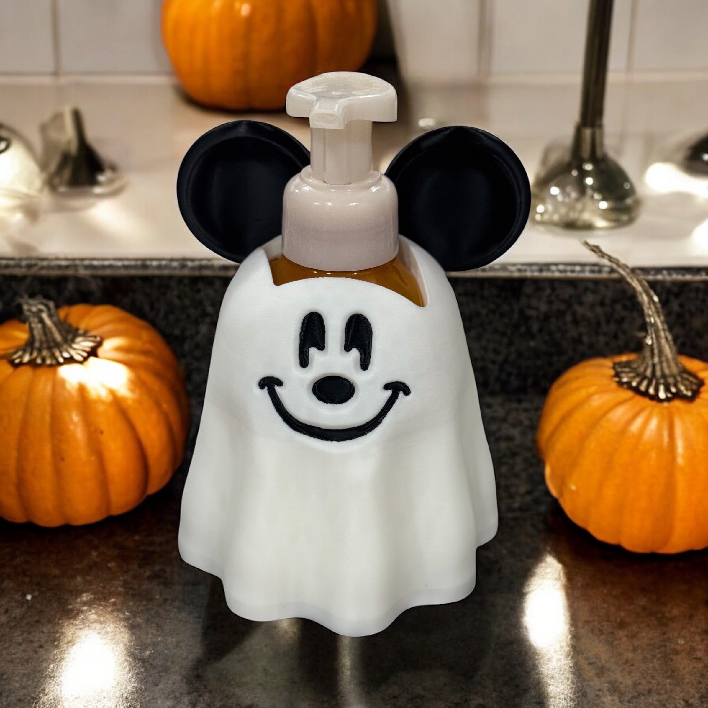 White ghost-shaped soap dispenser cover with mouse ears and a simple smiling face, standing between two pumpkins