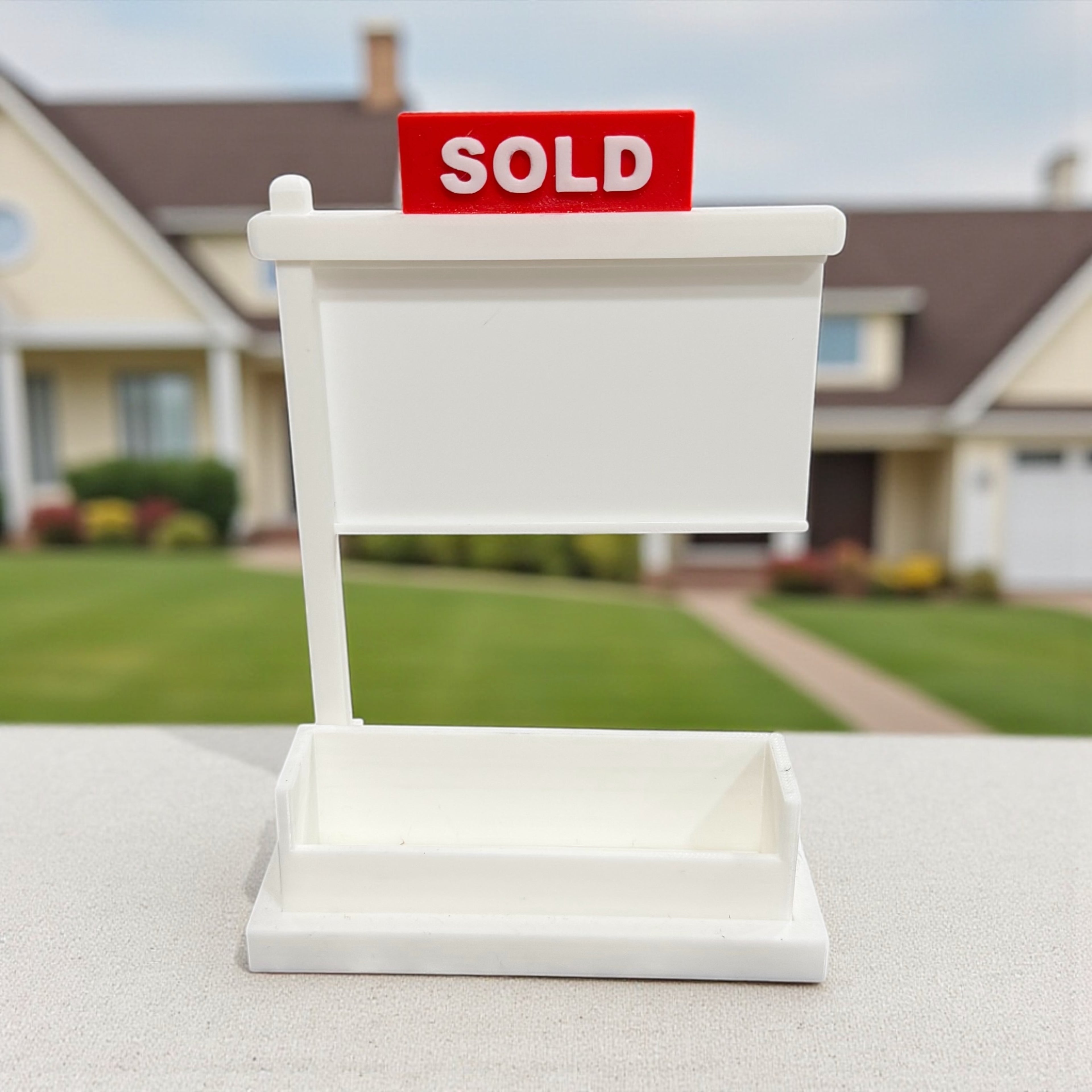 White miniature real estate yard sign with a SOLD rider, used as a desktop business card holder, photographed outdoors in front of a house