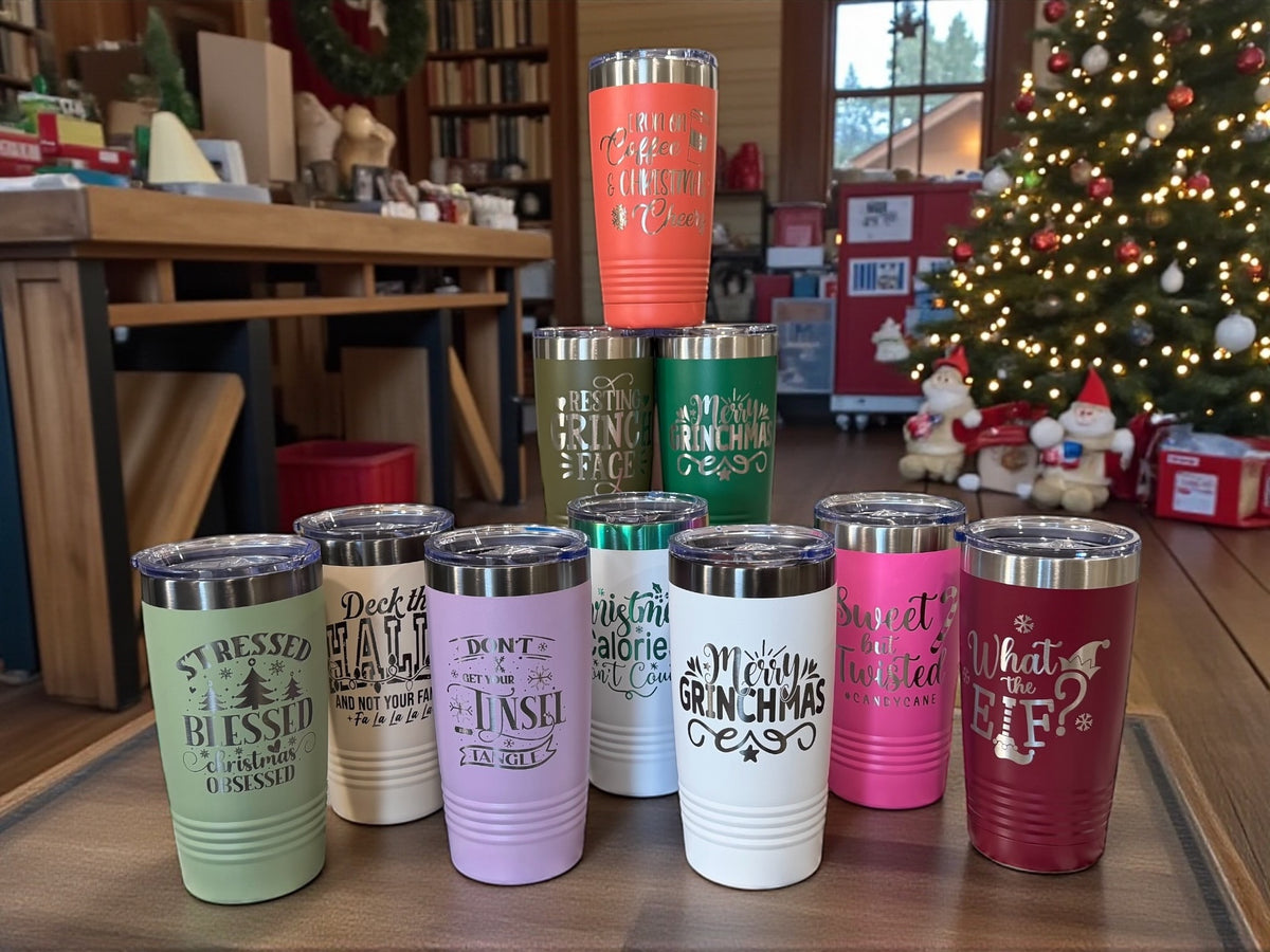 Eight engraved Christmas tumblers in assorted colors lined up in front of a decorated tree