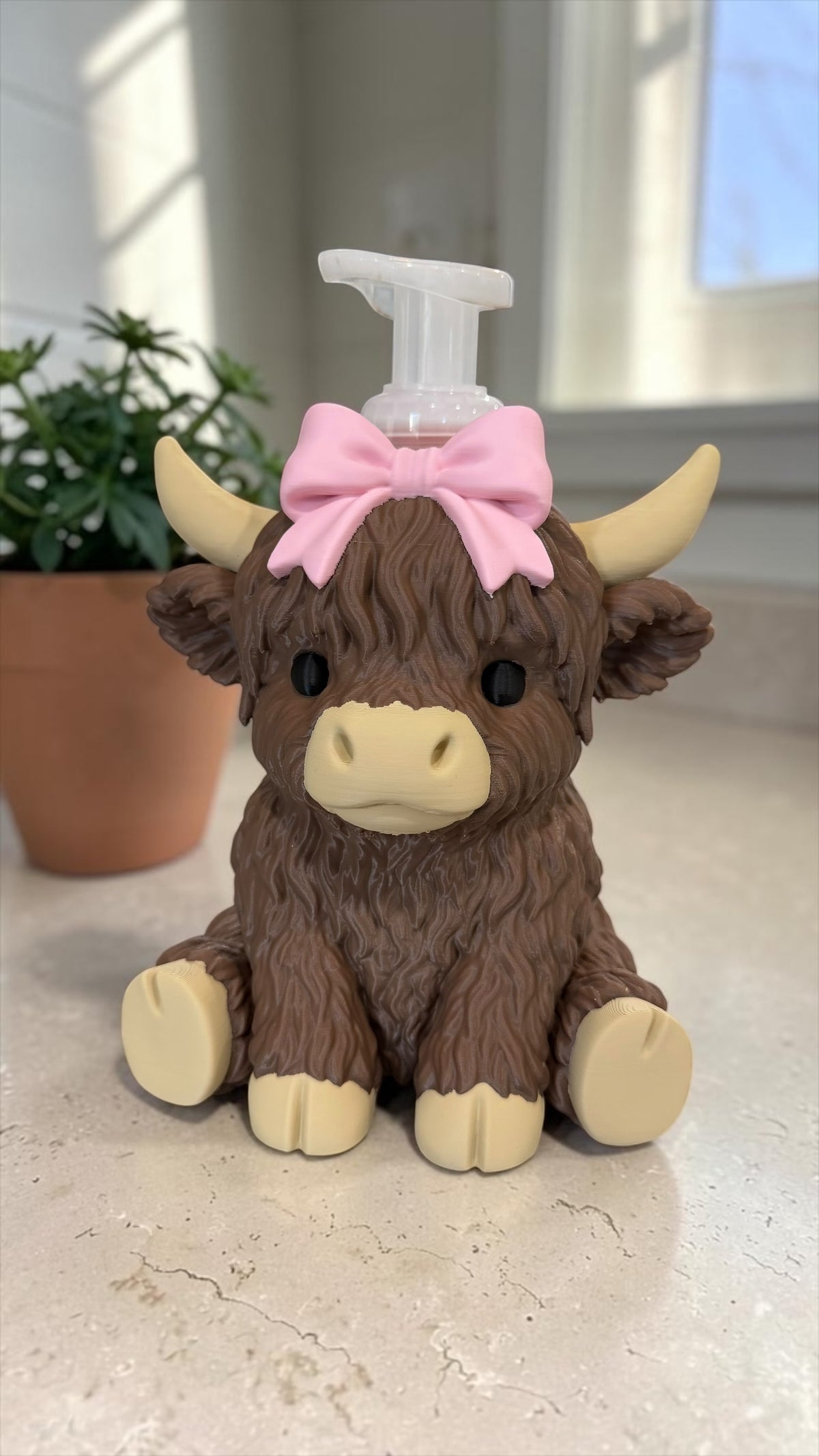 Highland cow soap holder with a pink bow, front view, with a foaming soap pump peeking out the top on a kitchen counter