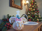 Gingerbread-styled soap dispenser cover with mouse ears, shown on a bathroom counter with a Christmas tree behind