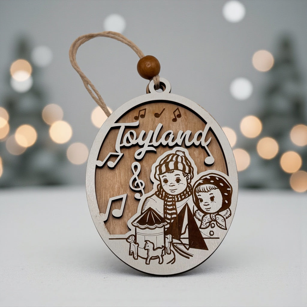 Wooden oval ornament engraved with a Toyland scene of two children and a toy drum, hanging against a snowy backdrop with soft white lights