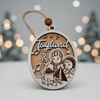 Wooden oval ornament engraved with a Toyland scene of two children and a toy drum, hanging against a snowy backdrop with soft white lights