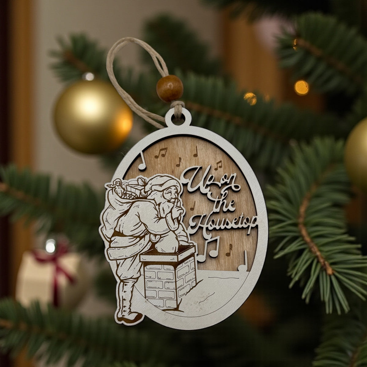 Wooden oval ornament engraved with an Up on the Housetop scene, hanging against a pine bough with gold baubles