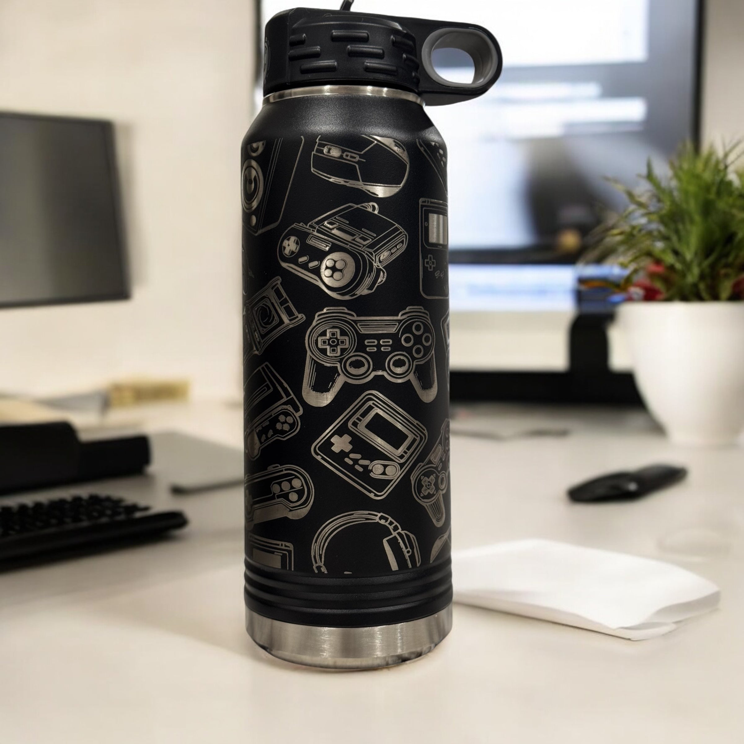 Black insulated water bottle engraved with a collage of video game icons, standing on a desk beside a keyboard and monitor