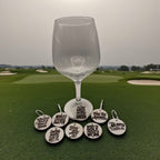 A wine glass on a golf course with a set of round wooden golf-themed wine charms laid out below it