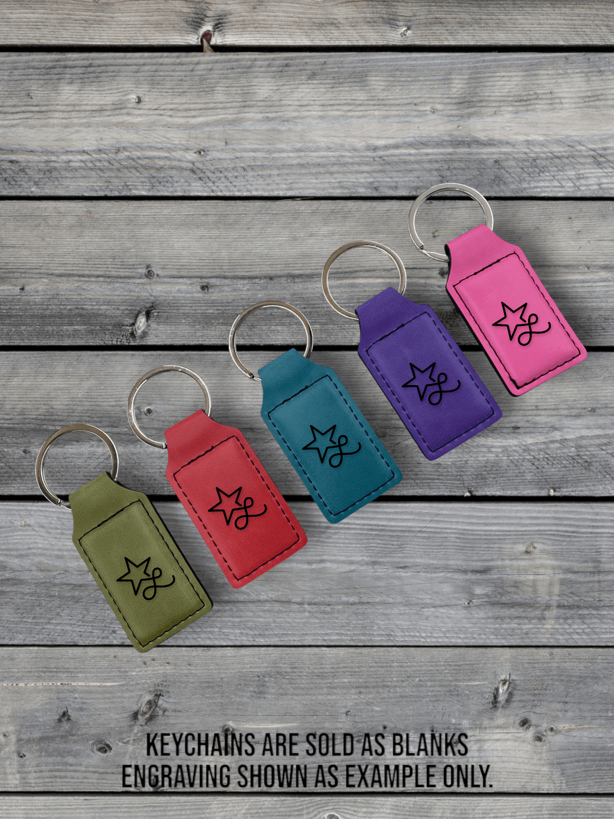 Five Premium Leatherette keychains in olive, red, blue, purple and pink, laid on weathered grey wood, each laser engraved with a sample design.
