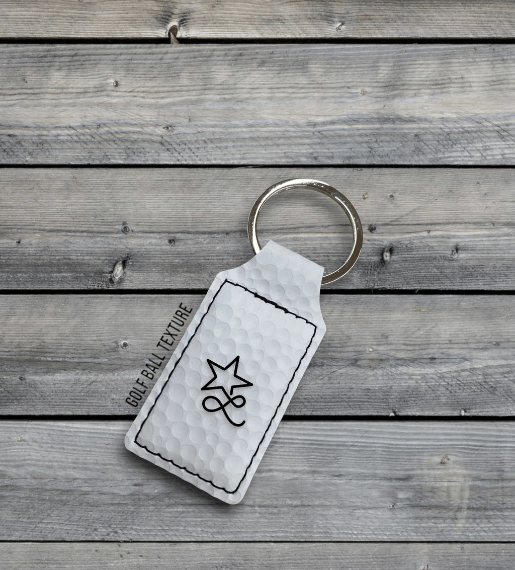 A white golf ball texture Premium Leatherette keychain with a split ring, lying on weathered grey wood, laser engraved with a sample design.