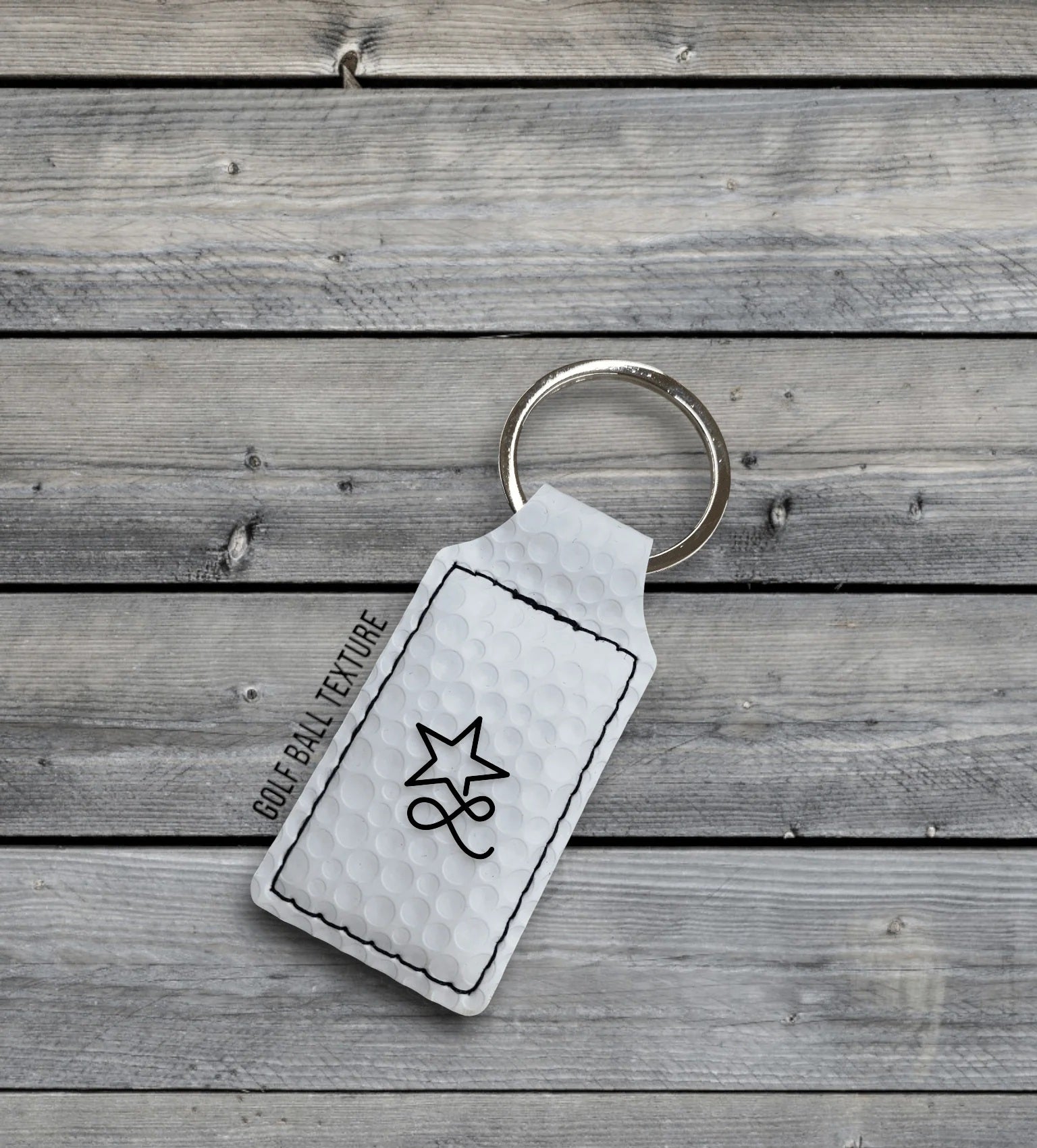 A white golf ball texture Premium Leatherette keychain with a split ring, lying on weathered grey wood, laser engraved with a sample design.