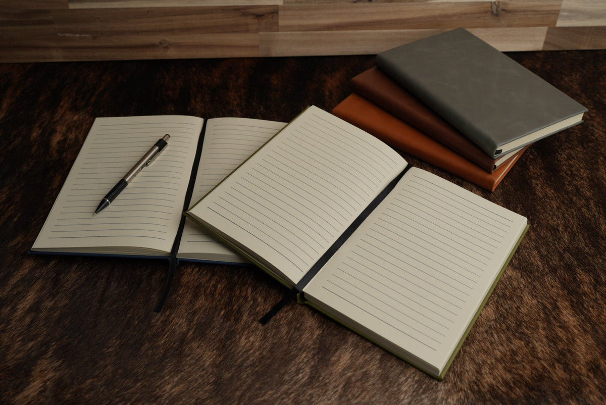 Two leatherette journals lying open side by side showing their cream lined pages, one with a pen across it, on a cowhide surface.