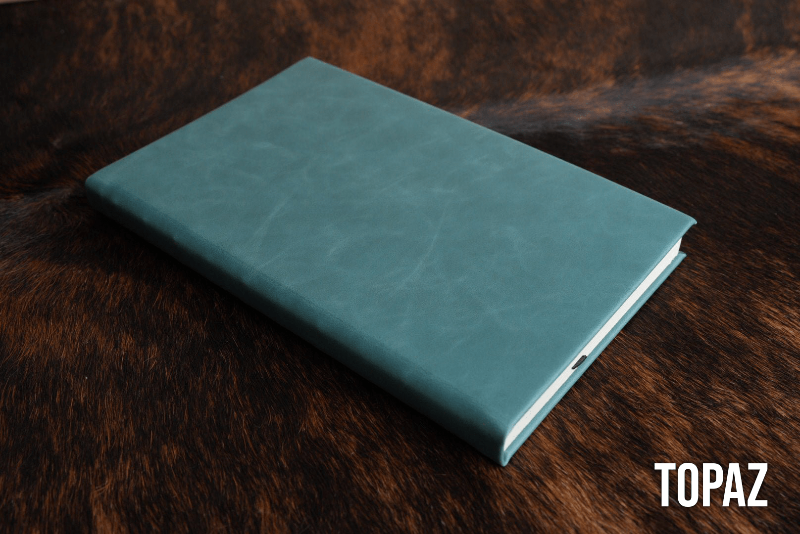 Topaz blue Premium Leatherette journal cover, shown closed on a cowhide surface
