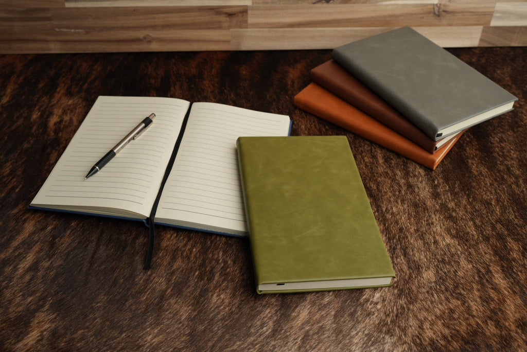 Leatherette journal lying open to show its cream lined pages, with a pen resting in the gutter and two closed journals stacked behind it.