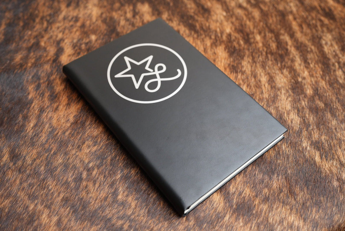 Black Premium Leatherette journal cover shown closed on a cowhide surface, with a sample logo engraved to reveal the silver layer beneath