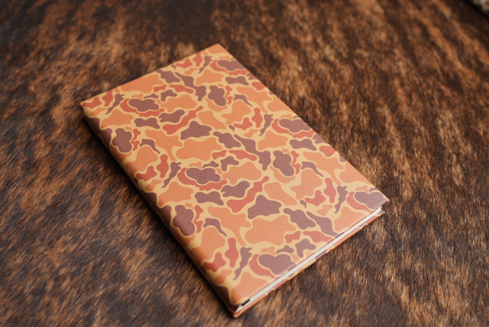 Drake Camo Premium Leatherette journal cover in orange, rust and tan, shown closed on a cowhide surface