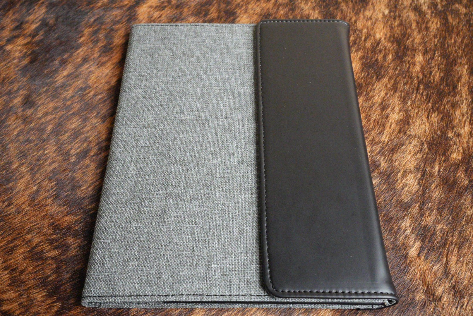 Black Premium Leatherette padfolio with a gray interior that engraves to silver, shown closed on a cowhide surface beside another colorway