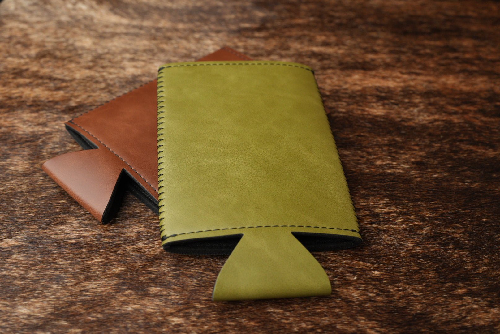 Brown and olive Premium Leatherette slim can coolers lying collapsed on a weathered wood surface.