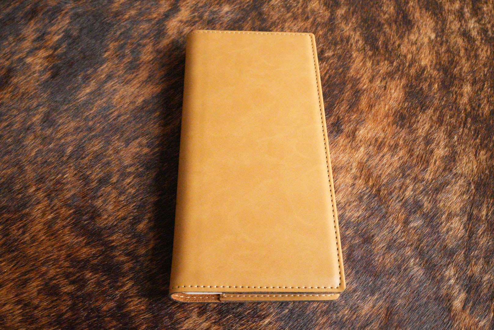 Saddle Tan Premium Leatherette tally book, shown closed on a cowhide surface
