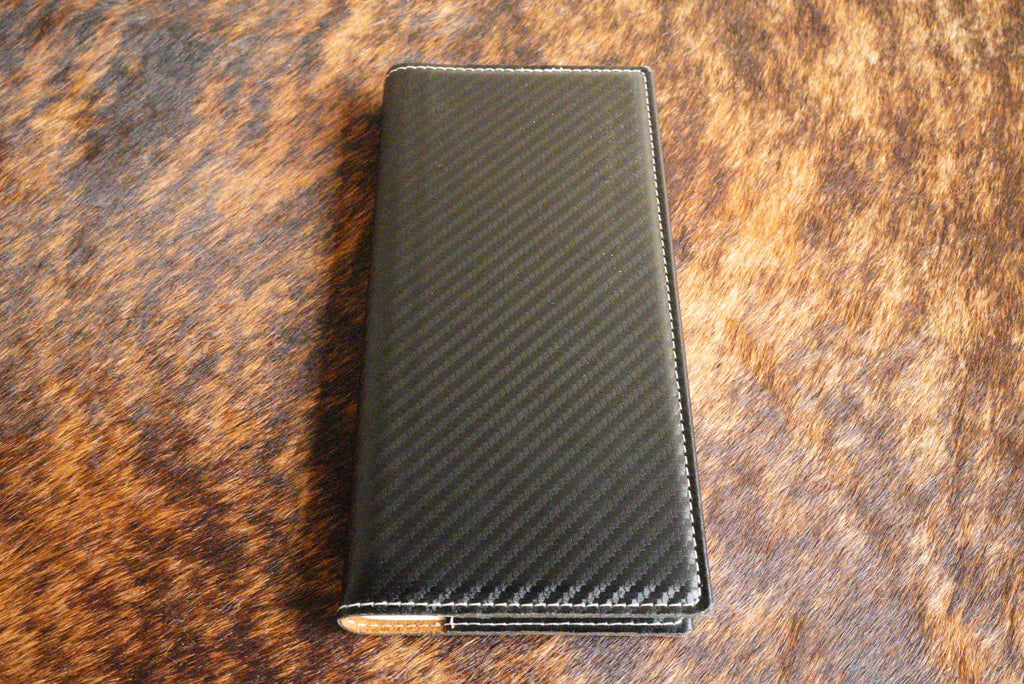 Black carbon fiber Premium Leatherette tally book that engraves to silver, shown closed on a cowhide surface