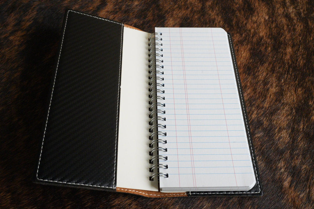 Open tally book showing the black carbon fiber inner cover, tan leather spine and spiral-bound lined notepad, resting on a cowhide surface.