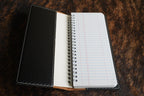Open tally book showing the black carbon fiber inner cover, tan leather spine and spiral-bound lined notepad, resting on a cowhide surface.
