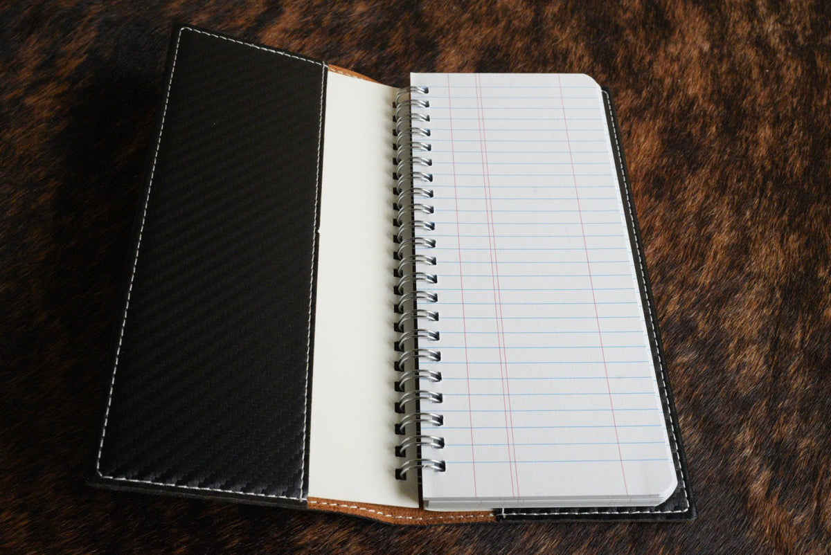 Open tally book showing the black carbon fiber inner cover, tan leather spine and spiral-bound lined notepad, resting on a cowhide surface.