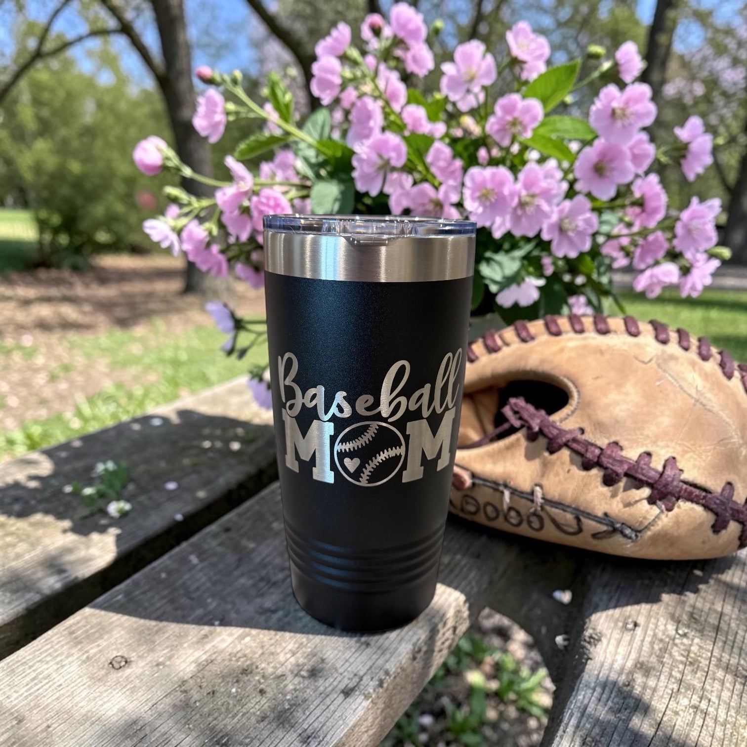 Black tumbler engraved with a Baseball Mom script design, staged on a counter with flowers and a serving board